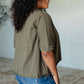 Model showcasing the Less Than Stressed Asymmetrical Distressed Top in olive green, featuring a unique design and relaxed fit.