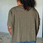 Model showcasing the back of a distressed asymmetrical top with paint splatter details in an olive green color.