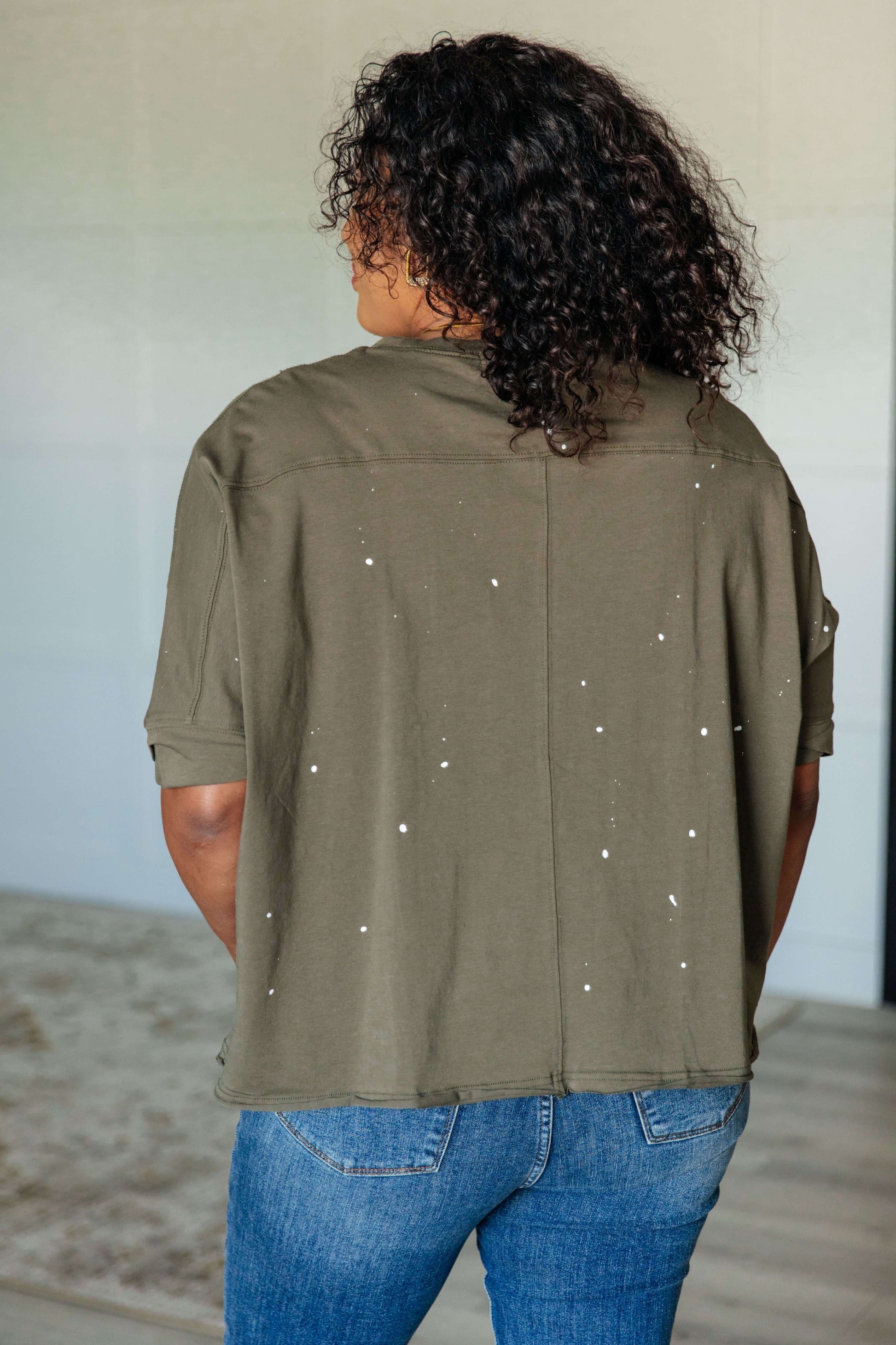 Model showcasing the back of a distressed asymmetrical top with paint splatter details in an olive green color.