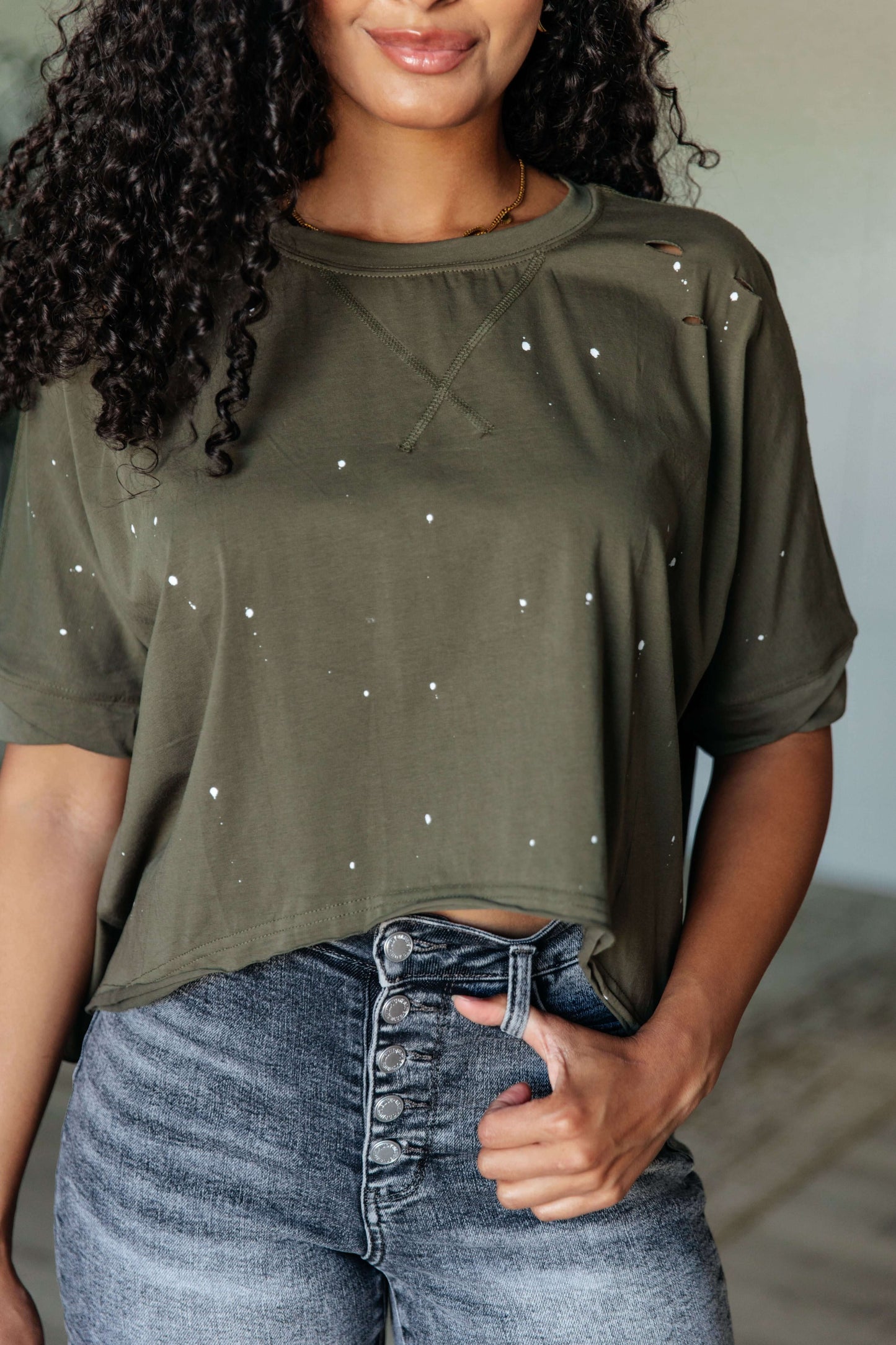 Woman wearing a distressed olive green asymmetrical top with paint splatter details and relaxed fit.