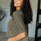 Woman with curly hair wearing an olive green asymmetrical distressed top, showcasing a stylish and edgy look.