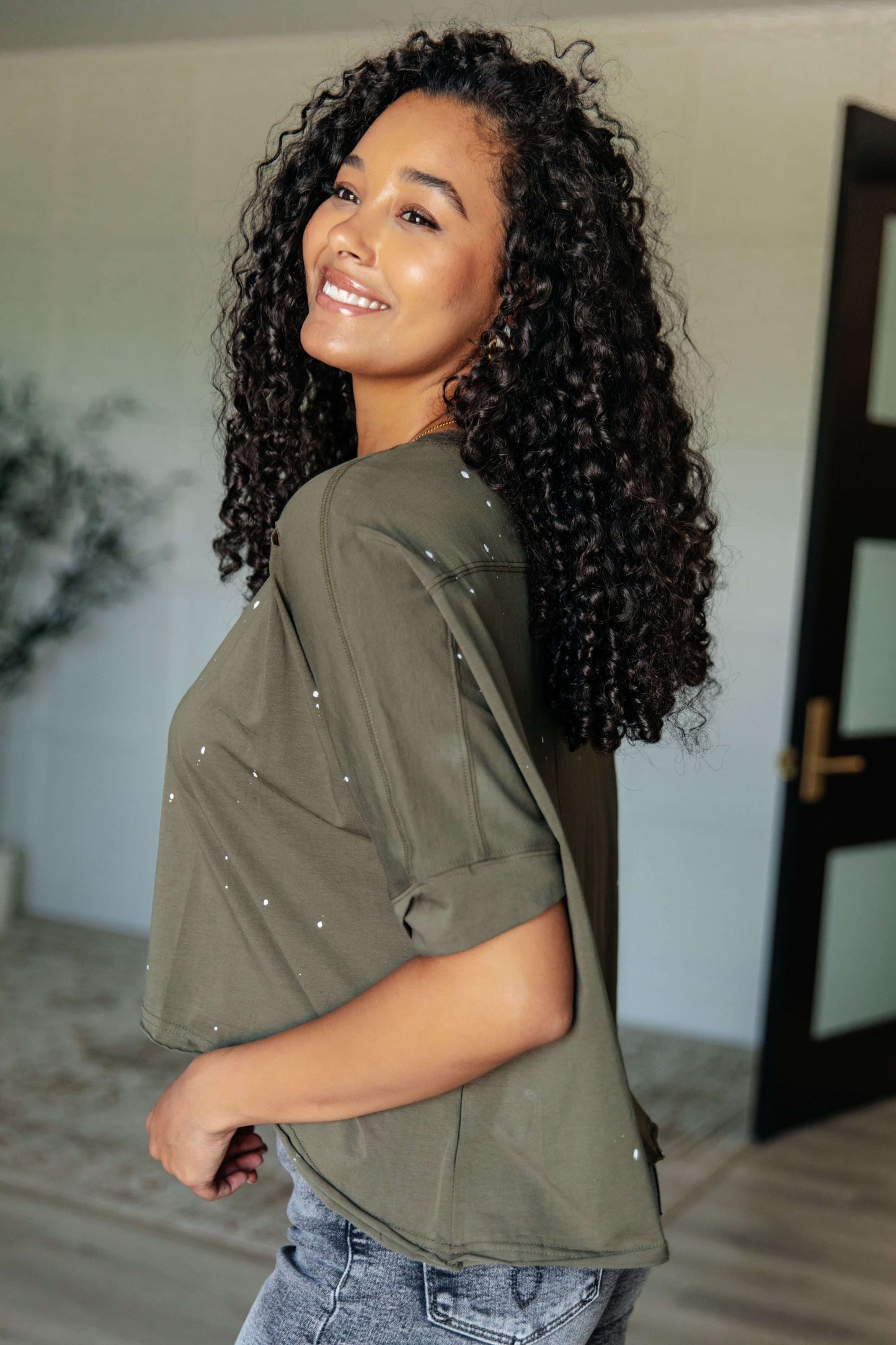 Woman with curly hair wearing an olive green asymmetrical distressed top, showcasing a stylish and edgy look.