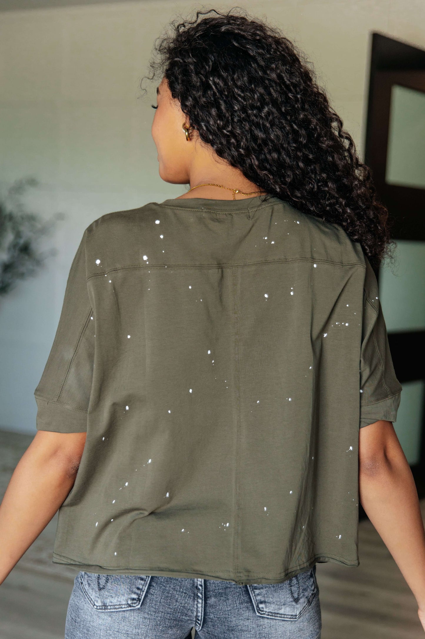 Back view of a woman in a distressed green top with paint splatter details and short sleeves, showcasing an asymmetrical hemline.
