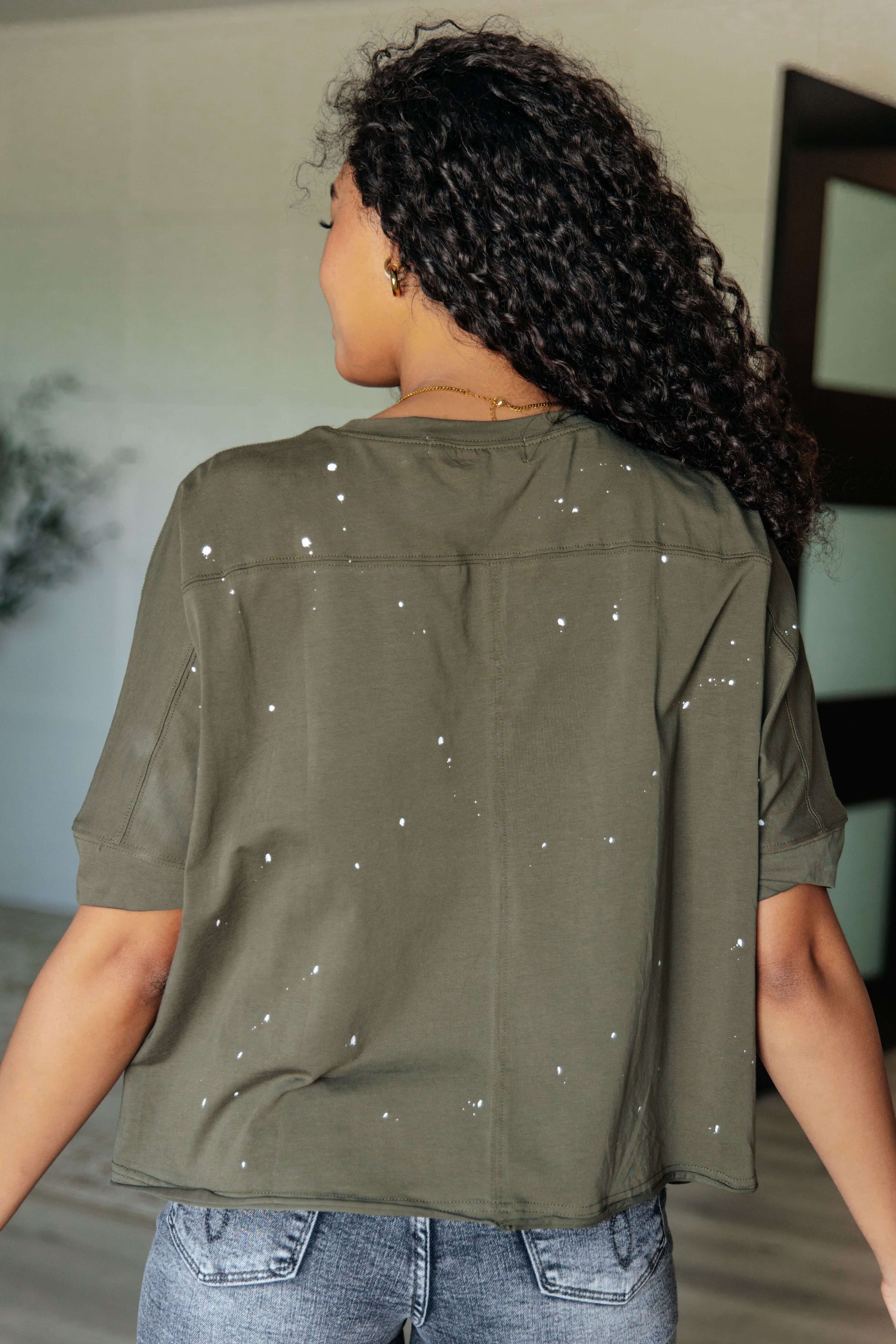 Back view of a woman in a distressed green top with paint splatter details and short sleeves, showcasing an asymmetrical hemline.